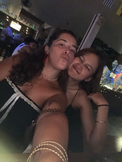 This is a selfie taken by two women at night, possibly in a bar or club in Khan Daun Penh, Cambodia. The woman in the foreground is making a kissy face at the camera, while the woman behind her has her eyes closed and a playful expression. Both women are wearing dark clothing. The background is blurred, but appears to be a lively scene with lights and other people. There is some text visible on a sign in the background, which appears to be in English, but it is too blurry to read clearly. The overall mood of the image is fun and festive.