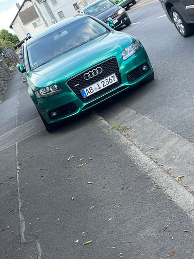 The image shows the front of a green Audi R8 parked on a street in Aschaffenburg, Germany. The car is facing towards the right of the frame, with its headlights and grill clearly visible. The license plate reads "AB 12367". It appears to be daytime, and the weather is fair. The street is paved with asphalt and has a curb made of concrete. There are some fallen leaves and debris on the ground next to the curb. In the background, there are some buildings and trees, suggesting an urban or suburban setting. The image is taken from a slightly elevated angle, looking down at the car. The car appears to be the main subject of the image. There are no people visible in the frame.