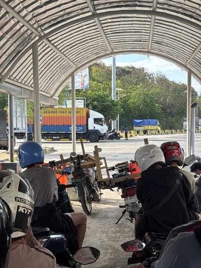 The image depicts a queue of motorcycles and a truck at a location resembling a port or ferry terminal in Upt Tambak Sari, Indonesia. A covered structure with a corrugated metal roof and metal beams extends over the scene, suggesting it might be a waiting area or loading bay.

In the foreground, three individuals are seated on motorcycles, viewed from behind. The person on the left wears a blue helmet and a grey t-shirt. The person in the middle wears a white helmet and a black hoodie with a crossbody bag. The person on the right wears a red and black helmet. Their motorcycles are parked in a line, some with wooden structures attached.

In the background, a large white and orange truck with the text "WASHENG KEKE MANDIRI" is visible, along with other vehicles and trees. The environment appears to be outdoors, with a bright sky and sunlight casting shadows. The ground is paved. A sign with the word "ZONA" and a cartoon deer can be seen on a pole. The presence of multiple vehicles, especially motorcycles, suggests this is a point of transit.