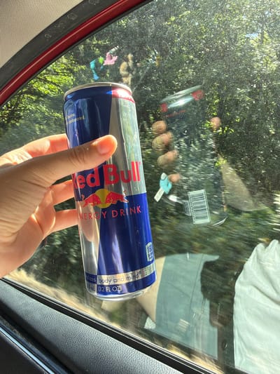 A person's hand is holding a Red Bull energy drink can in front of a car window, obscuring a portion of the view of trees and foliage outside. The can is blue and red with the Red Bull logo prominently displayed. The hand is fair-skinned and appears to be reaching towards the can. The background through the window shows lush green trees, suggesting an outdoor setting, possibly during daylight hours. There are some reflections visible on the car window, including what appears to be a barcode and possibly keys. The interior of the car is partially visible, with a dark-colored car door frame in the foreground. The image does not provide any specific information about the city of Seaside, United States.