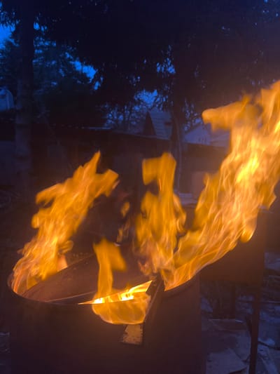 The image displays a vibrant, active fire burning in a dark outdoor setting, likely at night or dusk. Large, bright orange and yellow flames dominate the right side and center of the frame, swirling and extending upwards. These dynamic flames appear to be emanating from a dark, metallic object, possibly a fire pit or barrel, partially visible on the right.
The background is predominantly dark blue and black, indicating the time of day. Silhouetted against this dark sky are numerous trees and dense foliage, suggesting a natural or semi-natural environment such as a backyard or wooded area. Hints of a light-colored structure, possibly a building or fence, are faintly visible behind the flames and trees on the left side, reflecting some light. The image strongly emphasizes the intense glow and movement of the fire against the dark surroundings. No people or visible text are present in the scene.
