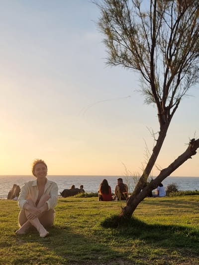 A woman is seated cross-legged on a grassy hill overlooking the sea at sunset in Palmachim, Israel. She is wearing a light-colored, open button-down shirt and appears to be nude from the waist down. Her arms are crossed in front of her, and she is smiling at the camera. In the background, several other people are visible, sitting on the grass and facing the sea. A barren tree with thin branches is positioned in the foreground, leaning to the right. The sky is a gradient of pale blue and warm orange, indicating dusk. Two thin, white contrails are visible in the sky above the horizon. The sea is calm and appears as a dark blue band.