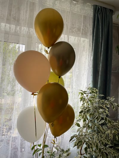 The image shows a group of seven balloons in front of a window covered with a sheer white curtain and a dark green curtain valance. To the right, a variegated leafy plant is visible. The balloons are a mix of colors, including gold, silver, peach, light green, and white. They appear to be tied together with gold ribbons. The location is not discernible from the image and cannot be identified as Ulukulevo, Russia.