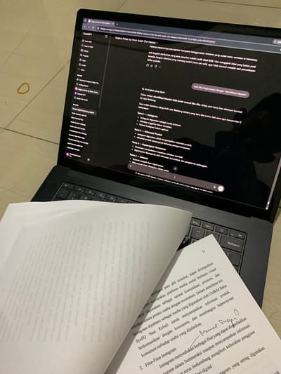 This media file captures a close-up, overhead view of a laptop and an open book, suggesting a study or work session. The primary objects are a sleek, dark laptop with its screen displaying a webpage, and a thick, white book with text and handwritten annotations. The laptop screen shows a website interface with text in Indonesian, including a URL referencing "chatgpt.com" and a title "Sarjana Pintar by Noor Asiah". Various menu items and content related to "ChatGPT" and "Skripsi Digital Marketing UMKM" are visible.

The book, lying open to a page with printed Indonesian text and a handwritten signature, is placed to the right of the laptop. A portion of the book's spine and several pages are visible, indicating it's a substantial read. The overall scene is likely indoors, given the artificial lighting implied by the illuminated laptop screen. There are no direct environmental cues like weather or time of day, but the focused activity suggests a dedicated work period. The presence of a yellow rubber band on the surface beneath the laptop suggests a casual, yet organized workspace. The overall mood is one of focused productivity.