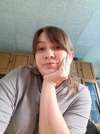 A medium shot photograph taken from a high angle captures a young woman with light brown hair and bangs resting her chin on her left hand, her gaze directed towards the camera. She is wearing a plain grey t-shirt. The background consists of a light blue ceiling with an embossed patterned texture and wooden cabinets with dark knobs. To the right of the woman, a glass-fronted cabinet is visible, containing items on its shelves. The overall setting appears to be an indoor domestic environment, likely a kitchen or dining area, within the city of Каменное, Russia.