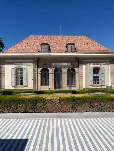 The image shows the exterior of the Villa du Parc in Cologny, Switzerland. The building features a red tile roof with dormer windows. The facade is made of light-colored stone and has several windows with gray shutters. There are glass panels in front of the building, creating an enclosed area. In front of the glass panels is a trimmed hedge. The ground in front of the hedge is paved with alternating gray and white stripes. The sky is clear and blue.