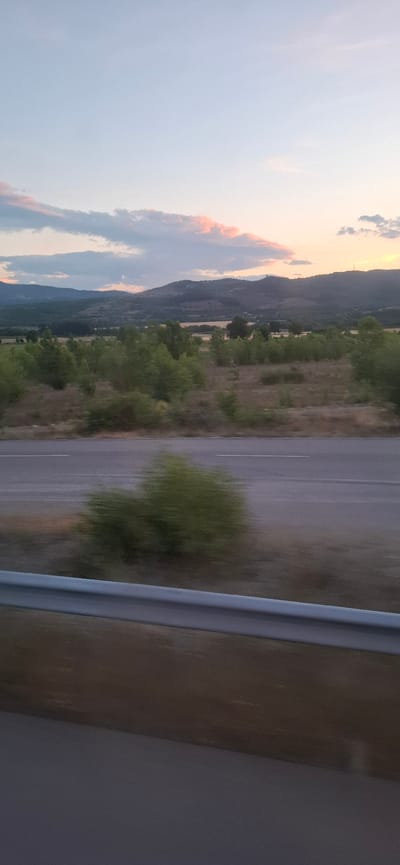 The image depicts a landscape scene with a road in the foreground, a guardrail, and a natural environment beyond. The sky is partly cloudy with the sun setting, casting a warm glow on the clouds and the distant hills. The ground is covered with dry vegetation and scattered trees. The road appears to be a highway, with white lines marking the lanes. There are no people or vehicles visible in the image. The overall impression is one of a serene, natural setting at dusk. The location is identified as being in the city of Mursalevo, Bulgaria.