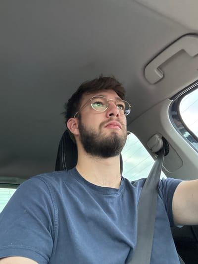 An image taken from within a vehicle shows a male subject seated, looking forward and slightly upward. The subject, appearing to be in his 20s or 30s, has short dark brown hair and a trimmed dark beard. He is wearing thin-framed, gold-colored, octagonal eyeglasses and a plain, dark blue short-sleeved t-shirt. A black seatbelt is visible across his left shoulder and chest. The interior of the vehicle features a light grey headliner, with a passenger-side grab handle visible on the right. The top portion of a dark car seat with light-colored stitching is behind the subject's head. Through the vehicle's window on the right, an overcast sky or bright exterior is visible, with indistinct background elements. The scene is situated in the city of Châtenois, France.