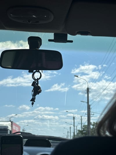 The image, taken from inside a vehicle, shows a view through the windshield. In the foreground, a rearview mirror hangs from the ceiling, adorned with a keychain featuring several charms, including a bell and what appears to be a small figurine. The windshield is partially obscured by the car's interior, including a dashcam mounted above the rearview mirror and a smartphone displaying a map application in the lower left corner. Outside, a bright blue sky with scattered white clouds dominates the scene. Several streetlights and power lines are visible along the side of the road. In the background, a blurred cityscape with buildings and a red truck can be seen. The scene appears to be captured in Usatove, Ukraine, based on the navigation display on the smartphone.
