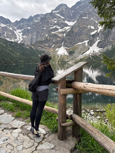 A woman in black leggings, grey shorts, a grey shirt, and a black jacket leans on a wooden fence overlooking a lake reflecting snowy mountains. The scene is in Brzegi, Poland. The ground is a stone path. The weather appears overcast.