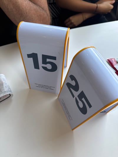 The photograph depicts a casual dining scene set on a white table, likely in San Buenaventura, Mexico. Two white, semi-cylindrical table locators with yellow trim are centrally placed, indicating order numbers for table service. The locator in the foreground displays the number "15," while the one positioned behind it shows "25." Both locators feature identical Spanish text: "Use este localizador para la entrega a la mesa. Ubicar en un lugar visible." (Use this locator for table delivery. Place in a visible spot.).

Partial views of two individuals are present in the image. On the far left, a person's arms are visible, one adorned with a dark bracelet. A portion of another person's leg is discernible in the bottom left corner. In the background, faint red and white packets, possibly condiments, can be seen. A small, rolled-up paper item, resembling a receipt or label, rests on the table in the bottom right corner. The bright and even lighting suggests an indoor setting during daytime or a well-lit interior. The scene captures a moment of waiting for an order at a restaurant or a similar establishment that utilizes table delivery systems.