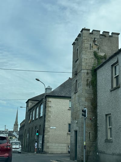 The image captures a street scene in Carlow, Ireland, with a focus on a stone tower that appears to be part of an old building, possibly a castle or historical structure. The tower is crenellated at the top and has ivy growing up its side. To the right of the tower, a gray, textured building with a window is visible. In front of the tower, a traffic light is mounted on a pole, showing a green light. A person in a white top and shorts stands near the traffic light. Behind the tower, another building with multiple arched windows is seen, and further in the distance, a church spire pierces the overcast sky. Cars are present on the street, including a red car in the foreground to the left and a white van behind it. A street sign reading "CHAFF ST" is attached to the gray building on the right. The overall atmosphere is subdued due to the cloudy sky.