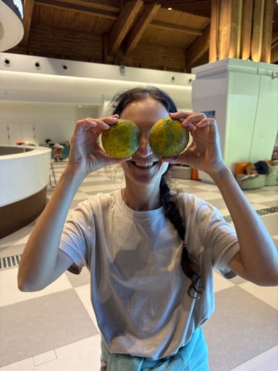 A woman stands in a modern, brightly lit indoor public space, likely an airport terminal or a large hotel lobby, situated in Tianya District, China. She occupies the center foreground, holding two round, green-yellow citrus-type fruits, each exhibiting dark surface blemishes, directly in front of her face, obscuring her eyes. She displays a wide smile with visible teeth. Her dark hair is secured in a long braid extending over her right shoulder. She wears a light-toned short-sleeved t-shirt and a light blue-green garment on her lower half. Her fingernails are manicured with light pink polish. The background features a high ceiling constructed with exposed dark wooden beams and lighter wooden panels, incorporating recessed circular lighting fixtures. The floor is tiled in a checkered pattern of alternating light grey and slightly darker grey squares. To the left, a curved white counter with a wooden trim is visible. To the right, a large white square pillar with vertical wooden slats on its upper section is illuminated, casting a warm glow. In the mid-ground, various lounge furniture pieces, including rounded green and orange seating pods, are present. One green pod is occupied by an individual lying prone. Additional blurred figures are visible in the background near the curved counter.