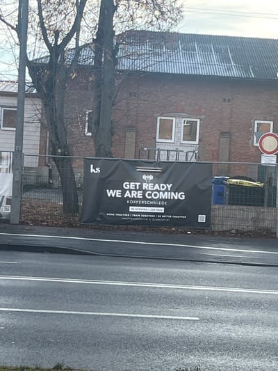 This image captures an outdoor street scene in Schweinfurt, Germany, dominated by a large black banner announcing an upcoming opening or re-opening. The banner, positioned prominently along what appears to be a main road, displays white text that reads: "GET READY WE ARE COMING" and "KÖRPERSCHMIEDE." Further details include "RE-OPENING / JAN 2024," along with a motto: "WORK TOGETHER / TRAIN TOGETHER / BE BETTER TOGETHER." The banner also specifies the address as "HAUPTBAHNHOFSTR 3 SCHWEINFURT." A small QR code is visible in the top right corner of the banner.

Behind the banner stands a two-story brick building with a corrugated roof, which appears to be undergoing renovation or awaiting it. Some windows on the building are visible, while others are covered or boarded up. A ladder leans against the building below one of the windows. High on the building, a red and white circular traffic sign (possibly a 'No Entry' sign) is attached. In the background, a bare-branched tree stands to the left, suggesting a cooler season like autumn or winter. The sky is overcast, indicating a grey, possibly cloudy, daytime. In the immediate foreground and to the right, a gray asphalt road with white lane markings is visible. A fence, a blue bin, and a yellow object can be seen behind the banner, next to the building. No people are visible in the scene.