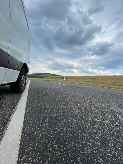 A white van is parked on the shoulder of a paved rural road. The van is white with a black trim and black tires. The road is asphalt with a white line marking the shoulder. A field of dry grass runs alongside the road, leading to a wooded hillside in the distance. The sky is cloudy, with patches of blue visible. The image appears to have been taken in the vicinity of Holzburg, Germany, a rural area with rolling hills and agricultural fields. There are no people visible in the image. The scene is still and quiet, suggesting the van is stopped for a break or an inspection.