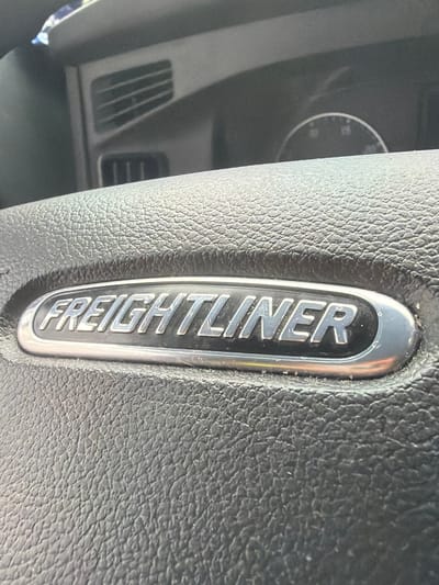 A close-up, eye-level, horizontal shot shows the "FREIGHTLINER" logo on the steering wheel of a truck. The logo is a silver oval with raised, capitalized lettering. The steering wheel is made of black, textured leather. In the background, out of focus, are parts of the truck's dashboard, including air vents and a circular gauge. The image appears to have been taken during the daytime, with bright lighting conditions. The location of this image is not specified in the provided data.