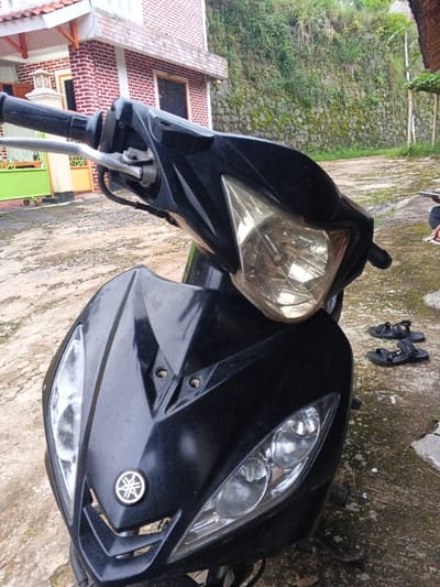 A black Yamaha scooter is parked on a mottled concrete or paved surface. The scooter, oriented towards the viewer, features a central main headlight with a yellowish tint and two clear, smaller headlights below, flanking a Yamaha emblem on the front panel. Two exposed screw holes are visible on the upper front fairing. To the left, a portion of a two-story building with red brick walls, white trim, and a terracotta-tiled roof is visible, with a green and orange gate structure further back. To the right of the scooter, a pair of black sandals lies on the ground, and a person's arm and leg are partially discernible, holding a white rectangular object. The background consists of a retaining wall heavily covered in green vegetation, indicating a hillside or embankment beyond, under an overcast sky. This scene is located in the city of Kyauk Hsoke, Myanmar.