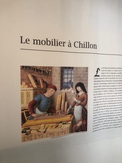 The image shows a printed page with the title "Le mobilier à Chillon" (The furniture at Chillon). The content refers to a collection of furniture at the Chillon Castle in Veytaux, Switzerland. It describes the collection's size, types of furniture (coffers, chests, seats, armoires), and historical periods represented. The text mentions the efforts of archaeologist Albert Naef starting in 1911 to complete the collection. The printed page includes an image of a man and woman working in a workshop. The text also discusses the significance of furniture as a source of information about history, savoir-faire, tastes, and social status.