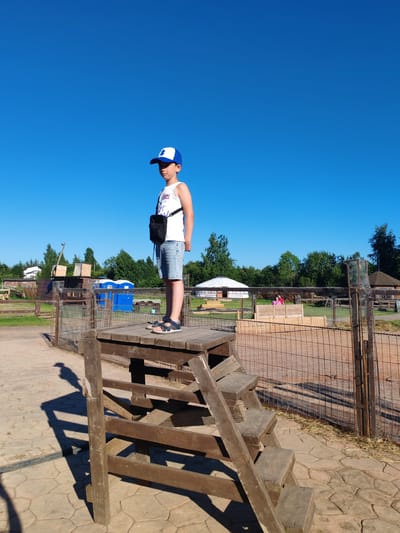 A young boy stands on a wooden platform at an outdoor petting zoo or farm in Lukashi, Russia. He is wearing a white tank top, denim shorts, a blue and white baseball cap, and sandals. He has a small black bag slung across his chest. The platform is elevated and has wooden steps leading up to it. The background shows a fenced-in area with various structures, including what appear to be animal enclosures, sheds, and possibly a yurt-like building. There are trees and a clear blue sky. The ground is paved with stone. The overall impression is of a sunny day at a rural recreational area.