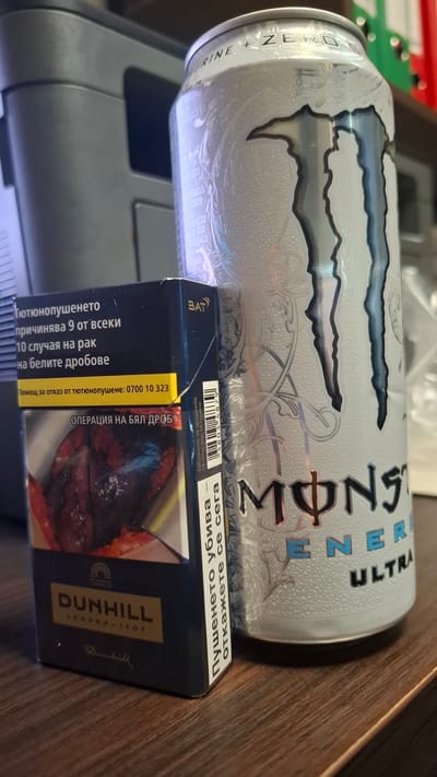 The image depicts a can of Monster Energy Ultra drink placed next to a pack of Dunhill cigarettes. The cigarette pack has Bulgarian text on it, including a health warning and a helpline number for quitting smoking. It is not possible to determine the location is Nikopol, Bulgaria from the image alone.