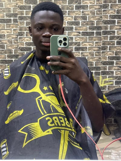 A dark-skinned man takes a selfie in a barber shop. He wears a black barber cape with yellow designs of scissors, combs, and stars. The phone he uses has a green case. The background is a textured wall. A pink hair-cutting machine cord is visible near his arm.