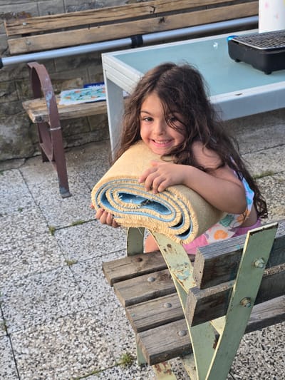 A young girl with long, dark, wavy hair is sitting on a wooden bench outdoors, holding a rolled-up beige rug with a blue lining. She is smiling broadly at the camera, revealing her teeth. The girl is wearing a pink and white patterned top and pink shorts. The bench has a weathered, rustic appearance with metal supports. In the background, a white table with a light blue top and a small black grill are visible. The ground is paved with a speckled gray material. The setting appears to be a patio or backyard area. The overall scene is bright and appears to be taken during the daytime.