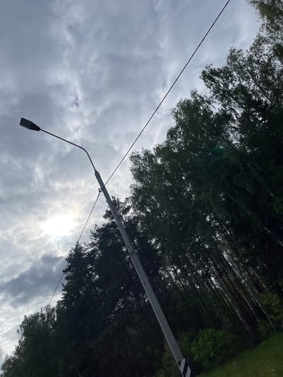 The image shows a street scene in Semyonovskoye, Russia, with a cloudy sky and sunlight breaking through. A street lamp extends from a utility pole, with power lines visible. Trees line the road, obscuring the view. A road sign is partially visible.