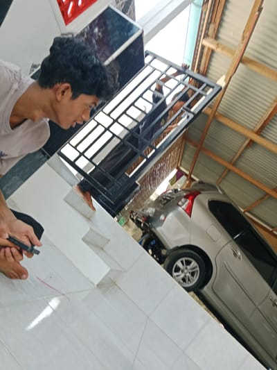 A view in Gampongjawa, Indonesia, from the ground level up at an angle. Two young men are holding a metal frame with a grid pattern over a white tiled area. One man is looking down at the tiles, while the other is up in the frame. A silver car is partially visible under a roof with wooden beams. A motorcycle is parked near the car.