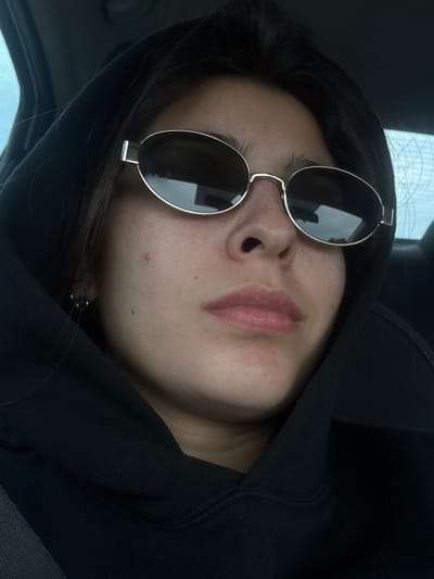 A person wearing a black hooded sweatshirt and round sunglasses is seen in a close-up shot. The person's face, including their nose and lips, is prominent. The background appears to be the interior of a car. The location is unclear.