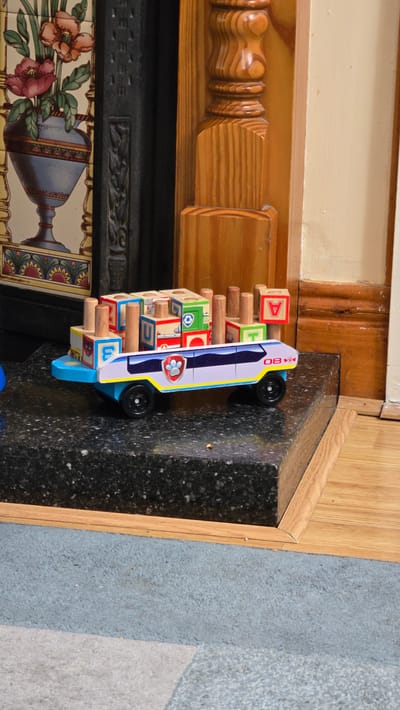 The image shows a toy bus with building blocks stacked on it. The bus is blue and white with a Paw Patrol logo and the text "M80" visible on its side. The building blocks are wooden and have letters, numbers, and symbols on them. Some of the blocks show the letter "A", the letter "U", and a Paw Patrol logo. The toy is positioned on a dark, speckled surface, which appears to be a countertop or mantelpiece, in front of a wooden structure with carved details. In the background, there is a wall with wooden paneling and a glimpse of decorative tiles on what might be a fireplace. The lighting suggests it is daytime. The overall scene appears to be indoors, possibly in a home setting in Kilkenny, Ireland. There are no people visible in the image.