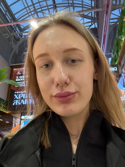 A selfie of a blonde woman with a nose piercing is the primary subject. She is wearing a black jacket, and a small heart tattoo is visible on her neck. The background shows a glass-roofed structure and signs with text, including "ЛЕКЦИ ХРИС ЖАНН," "3 ΑΠΗ 19:00," and "SKI." The context suggests an indoor location, possibly in Ol'ginskaya, Russia, given the Cyrillic text on the signs.