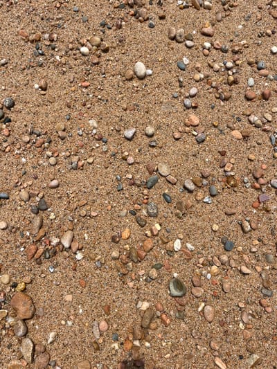 The image shows a close-up view of a sandy beach in Tamariu, Spain, at coordinates 41.918 latitude and 3.207 longitude. The sand is tan in color and is covered with numerous small and medium-sized pebbles of various shapes and colors. The pebbles are predominantly gray, brown, and white. There are no people or objects in the image other than the sand and pebbles. The image focuses on the texture and composition of the beach surface.  The scene suggests a calm and peaceful environment, typical of a beach setting.
