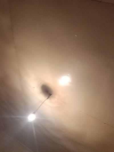 This is a dimly lit, high-angle indoor shot of a ceiling in Naray, Kyrgyzstan. The ceiling appears to be textured and off-white, with some visible streaks and smudges, suggesting it may be aged or in need of cleaning. Two bare light bulbs hang from wires, illuminating the space. The bulb on the right is brighter and appears to be closer to the camera, casting a strong glare and a shadow on the ceiling. The bulb on the left is also illuminated but less intensely. There is a faint, small, bright green speck of light in the upper right quadrant of the image, possibly a reflection or a lens flare. The overall impression is of a simple, functional lighting setup in a domestic or utilitarian indoor environment.