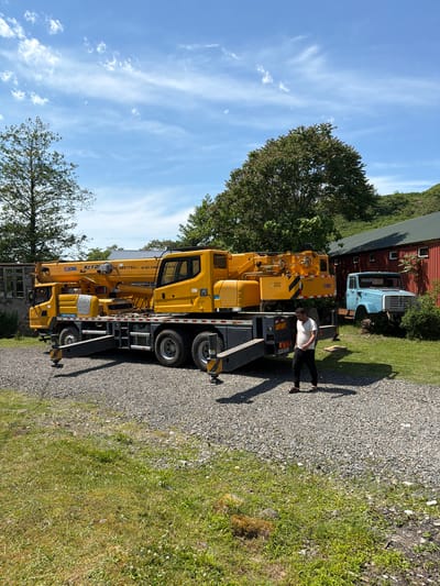 A yellow XCMG crane is parked on a gravel driveway in Kapreshumi, Georgia. A man in a white shirt and dark pants stands next to the crane. In the background, there is a red barn and a blue truck. The sky is blue with wispy clouds, and there are trees surrounding the area. The crane has black and yellow diagonal stripes. Patches of grass grow on the side of the driveway.