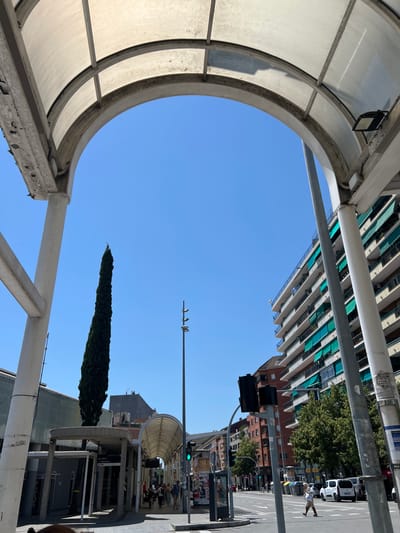 The image is taken from a low angle, looking up through a partially enclosed, semi-circular structure, possibly a bus stop or sheltered walkway, in Granollers, Spain. The sky is a clear, bright blue. In the background are buildings, including an apartment complex with numerous balconies and green awnings, and a taller brick building. Further back, figures of people are walking on the sidewalk. A tall, narrow cypress tree is visible. The perspective suggests the structure is designed to provide shelter while allowing an unobstructed view of the sky. Three birds are perched on a utility pole against the clear sky. In the distance, there is another structure identical to the one where the image was taken. A traffic light shows a green light.