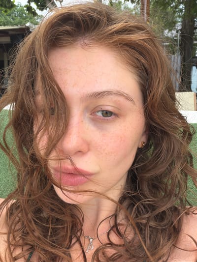 This media file is a close-up, rotated selfie of a young woman with long, wavy, light brown hair. Her face is partially obscured by her hair, but her left eye, nose, and lips are visible. She has light skin with numerous freckles scattered across her cheeks and nose. She is wearing a delicate silver necklace with a pendant shaped like the letter "P." A gold hoop earring is visible in her left ear. Her lips are closed and appear to be a natural pink color. The background is out of focus, but suggests an outdoor setting with green textured ground, possibly a towel or mat, and some greenery and structures visible in the distance. The lighting suggests it is daytime, and the overall mood is casual and relaxed. The location context provided is Ao Cho, Thailand, which is a coastal area.
