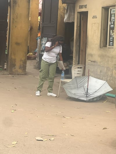 This image depicts a street scene in Ikotun, Nigeria. A female subject, with long dark hair, wearing a white t-shirt, olive green cargo pants, and white sneakers, stands on a dusty, light brown ground scattered with dried leaves. She holds a mobile phone to her right ear, appearing to be on a call, while simultaneously holding white documents or papers in her left hand. Sunglasses rest on her head.
To the right of the woman, a white plastic chair is positioned. A clear plastic water bottle rests on the seat of this chair. Adjacent to the chair, a large, grey umbrella lies partially inverted or collapsed on the ground, exposing its internal structure and a red central pole.
In the background, a light beige concrete building with a wooden door is visible. Above the door, the words "SECURITY POST" are painted in dark letters, with "ORGANIZED BY MULJEH EXCO" painted below. A window to the right of the door contains multiple small papers or photos. To the left of the female subject, a narrow opening or alleyway framed by a tall, faded yellow concrete pillar reveals glimpses of other structures and a bare human foot in the distance.