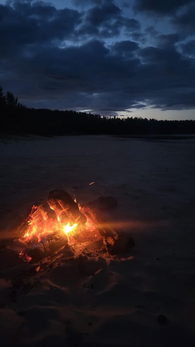 A campfire burns brightly on a sandy shore, illuminating logs of wood with vibrant orange and yellow flames. The sand dunes are dark and textured, with subtle shadows cast by the firelight. In the background, a dark line of trees stretches across the horizon, silhouetted against a cloudy twilight sky. The sky is a deep blue, with dramatic, heavy clouds that suggest the end of the day or approaching weather. A faint glow emanates from the horizon, hinting at the last vestiges of daylight. The overall scene conveys a sense of solitude and natural beauty.