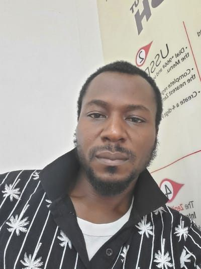 An adult male of African descent is depicted in an indoor setting, identified as Sabon Gari North, Nigeria. He is positioned from the upper torso up, looking directly at the camera with a neutral expression. He has short, dark hair, a trimmed dark beard, and a mustache. He is wearing a black collared shirt with white vertical stripes and white palm tree patterns, layered over a white t-shirt. The background consists of a plain white wall on the left. On the right, a vertically oriented banner or sign is visible, displaying promotional text and logos, some inverted or rotated. Identifiable elements on the banner include "USSD", "Dial *966#", "The Zenith Bank", and "Create a 6-digit PIN", along with red logos, indicative of a banking or financial service advertisement.