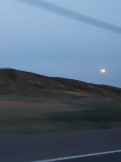 The image shows a blurred, low-light view of a landscape at dusk or dawn. A dark, undulating hillside occupies the middle ground, with patches of lighter vegetation. The sky above is a pale blue-gray, with a faint, indistinct cloud formation or contrail stretching across it. To the right of the hillside, a small, glowing, round object is visible in the sky, possibly the moon or a distant light. The foreground is dominated by a blurred dark surface, likely a road, with a faint white line visible near the bottom edge, indicating a lane marker. The overall impression is one of motion blur, suggesting the photo was taken from a moving vehicle. The location is identified as Нарын ауылдық округі, Kazakhstan.