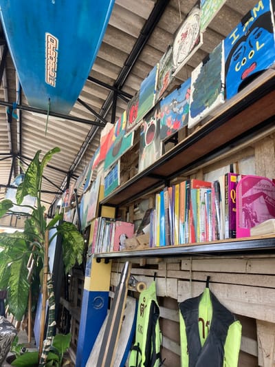 This media file captures a vibrant and eclectic indoor or covered outdoor space, likely in Surat Thani, Thailand. The scene is filled with a variety of objects, creating a bohemian and artistic atmosphere.
Dominating the upper and middle sections are numerous colorful painted wooden boards of various sizes, suspended from the ceiling. These boards display diverse artwork, including a blue one with a cartoon face and the text "I'M COOL," abstract designs, portraits, and scenic depictions. They hang above a blue surfboard with the clear text "OLEBOARDS" and partially visible "SURFBOARDS."
To the right, a large wooden bookshelf is packed with books, some identifiable by their English titles like "Quit Your Job" and "Queen," alongside many volumes with Thai script on their spines. Above the books, a pink mesh organizer is visible. The shelving unit itself appears to be constructed from or decorated with wooden pallets, contributing to a rustic or upcycled aesthetic.
Below the bookshelf, various sporting and water-related equipment is stored. Two lime green and black life jackets, one partially revealing the brand "BILLABONG," are visible, alongside multiple longboards or skateboards. The background features a corrugated metal roof, suggesting a sheltered area open to the elements or a simple construction style.
A large plant with variegated green and light green leaves adds a touch of nature, its foliage visible in the lower right portion of the image. The overall impression is one of a relaxed, creative, and possibly recreational space, such as a surf shop, cafe, or community hub, where art, literature, and water sports converge. No direct human activity is shown, but the arrangement implies a space for leisure and engagement. The time of day appears to be daytime, given the natural light.