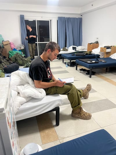The image shows a young man sitting on a bed in a room in Beersheba, Israel (31.249, 34.782). The room appears to be a dormitory or barracks, with multiple beds lined up along the wall. The young man is wearing a black t-shirt and olive green pants. He is writing in a notebook with a blue pen. There are other men in the room, one of whom is standing by a window. There are several beds with white linens and blankets in the room. The floor is tiled. There are windows, curtains, and a door in the room. The man is seated on the bed, with his feet resting on the floor. The bed is made of metal and has a white comforter. The man is looking down at the notebook in his lap.
