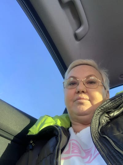 An adult female is depicted in a close-up, upward-angled portrait taken from within a vehicle in Gol'yany, Russia. She has short, light-colored hair and wears glasses with thin, gold-colored frames. She is dressed in a dark, possibly black, puffer jacket with a visible zipper, worn over a white top featuring a partially visible light pink graphic. A bright neon green garment or lining is visible around her neck area, protruding from the jacket. The subject maintains a neutral expression, looking directly at the camera. Above the subject, a clear blue sky is visible, suggesting an open sunroof or window. The interior of the vehicle shows a light-colored (beige/gray) headliner and a matching overhead grab handle.