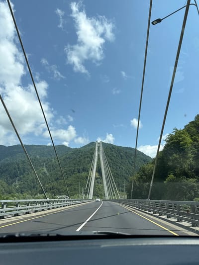 The image, taken from the perspective of a vehicle's dashboard, shows a view of the road and the surrounding landscape from a bridge. The bridge is a modern cable-stayed bridge with a wide asphalt road surface, marked with white and yellow lines. The bridge has metal railings on both sides. In the distance, on the road, a white car is visible. The bridge spans across a valley, with lush green mountains on either side. The sky is blue with scattered white clouds. A street lamp is visible on the right side of the bridge, attached to a support cable. The landscape suggests a rural or mountainous area. The context provided by "Kepsha, Russia" places this scene in the vicinity of the resort village of Kepsha, located in the Lazarevsky District of the city of Sochi, Krasnodar Krai, Russia, which is known for its mountainous terrain and proximity to the Black Sea. The bridge itself is likely part of the infrastructure development in the region, possibly related to tourism or transportation.