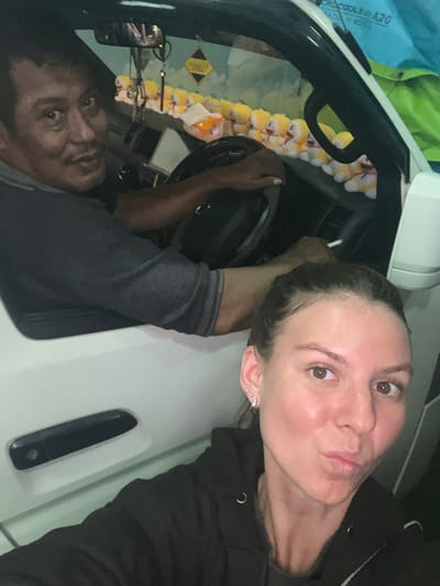 The image shows a selfie taken inside a white vehicle, possibly a van, with a man in the driver's seat and a woman in the foreground. The man, with dark skin and hair, is looking towards the camera with a slight smile. He is wearing a dark shirt and has a cigarette in his hand, which he appears to be smoking. The interior of the vehicle is decorated with numerous small yellow stuffed toys, which are arranged along the dashboard and windshield. A small yellow diamond-shaped sign is also visible hanging from the rearview mirror.
The woman in the foreground, with fair skin and brown eyes, is also looking at the camera and making a kissing gesture with her lips. She is wearing a black hooded jacket. The environment appears to be indoors or at night, given the lighting. There is no specific visual information to confirm the location in Gunung Malang, Indonesia, but the presence of the man with Asian features and the general context suggest an Asian setting. The scene depicts a casual moment shared by two individuals inside a vehicle.