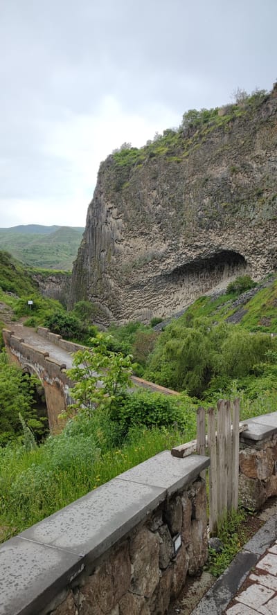 The image depicts a landscape in Garni, Armenia, featuring a bridge and a striking rock formation known as the "Symphony of Stones". The bridge, made of stone or concrete, spans a deep gorge filled with lush greenery. The rock formation is a massive cliff face characterized by its unique basalt columns, giving it an organ-like appearance. A prominent cave-like indentation is visible within the cliff face. The foreground includes a stone wall with a concrete top and a wooden fence post. The sky is overcast, suggesting cloudy weather conditions.