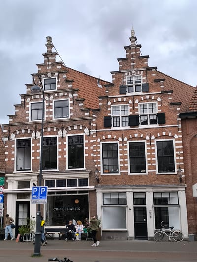 The image captures a street view in Haarlem, Netherlands, featuring two traditional Dutch canal houses. The buildings are constructed with red brick and accented with white decorative elements and stepped gables. The house on the left has a shopfront with the name "COFFEE HABITS" visible in the window. Several people are present: some sitting on benches outside the coffee shop, others walking by. A lamppost stands prominently in front of the coffee shop. A white bicycle is parked to the right of the frame, next to a shop with large windows. The sky is overcast, indicating a cloudy day.