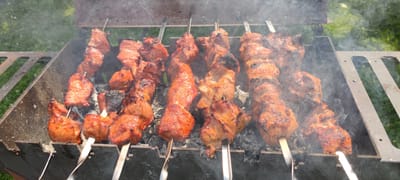 The image shows skewers of meat grilling over charcoal in a metal brazier, outdoors. The meat appears to be pork, cut into cubes and seasoned, with some pieces showing charring from the heat. Smoke rises from the grill, indicating the cooking process is underway. The brazier is placed on a surface of grey ash, with the meat positioned above the hot coals. In the background, blurred green grass and foliage suggest a natural or park-like setting. The overall scene depicts a casual outdoor cooking event, likely a barbecue or picnic. The image does not contain any individuals or specific landmarks that would allow for precise location identification within Пацейкаўскі сельскі Савет, Belarus.