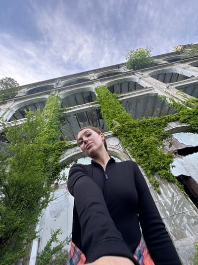 A low-angle selfie taken by a light-skinned individual with brown hair, against the backdrop of an overgrown architectural structure. The individual is wearing a black zip-up jacket and a multi-colored garment. The structure features a series of archways, some covered in dense green vegetation, including ivy and trees. The sky is visible in the upper left corner, displaying a cloudy, overcast appearance. The architectural style suggests a historical building in disrepair. Given the location details provided, it is presumed that the image was taken at an abandoned structure in the city of Tsanbaikwara, Abkhazia.