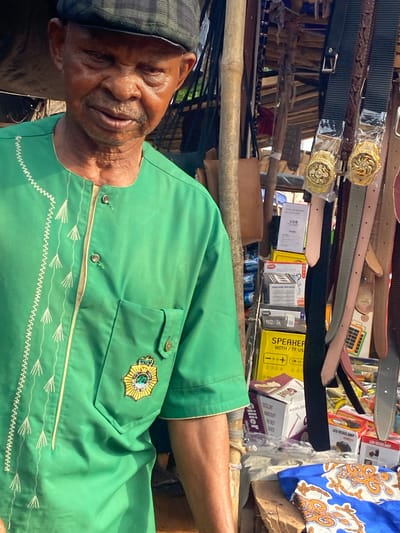 The scene depicts an elderly man positioned in front of what appears to be a market stall in Amaseri, Nigeria. The man, of dark complexion, wears a dark, patterned flat cap and a vibrant green tunic with a stand-up collar. The tunic features intricate white embroidery down the left front and a single pocket on the left chest, adorned with a golden, shield-shaped emblem displaying a crown motif. His head is slightly tilted downward, and his facial expression is neutral.
To the right of the man, numerous belts are suspended vertically, including black, light pink, and grey varieties. Two black belts feature prominent, ornate gold-colored buckles enclosed in clear plastic packaging. Below the hanging belts, various retail boxes are stacked. Visible labels on these boxes indicate "SPEAKER WITH / TF US," "WIRELESS SPEAKER," "LED HEADLAMP," and a white paper sign mentioning "USB." A blue fabric with an orange and white paisley-like pattern rests on a wooden surface in the lower right foreground. A rustic wooden pole, resembling bamboo, serves as a structural element in the mid-ground on the left side of the frame. The scene is illuminated by bright daylight.